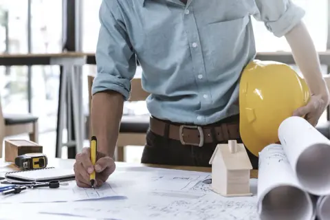 Ein Bauleiter, der als Generalunternehmer Baupläne prüft. Er hält einen gelben Helm und einen Stift.