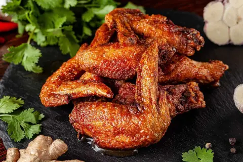 A pile of golden-brown, crispy alette di pollo, glistening with sauce, are served on a dark slate. Coriander, ginger, garlic, and peppercorns complete the appetizing presentation.