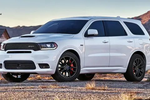 Biały Dodge Durango 3.6 SRT z czarnymi felgami i czerwonymi zaciskami hamulcowymi stoi na żwirowej drodze, w tle góry i opuszczony dom.