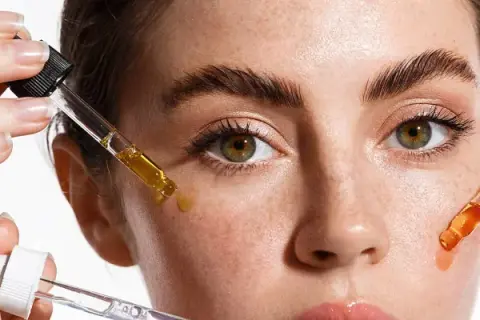 Hände tragen Pipetten mit Seren auf das Gesicht einer Frau auf. Diese Seren enthalten oft Peptide, die für die Hautpflege wichtig sind.