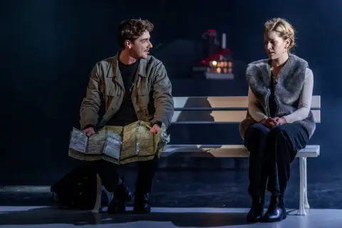 Para na ławce, mężczyzna z mapą, kobieta w futrzanej kamizelce. Scena z dramatu "Jelegnia Góra" pełna napięcia.