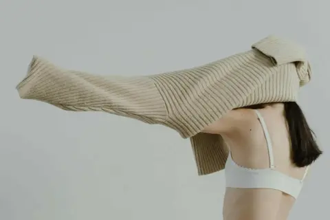 Eine Frau mit dunklem Haar trägt einen weißen BH. Ein beiger Strickpullover wird über ihren Kopf gezogen.