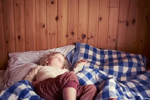 En 3-åring sover i en säng med rutiga täcken. Det är vanligt att en 3-åring kissar på sig på natten.