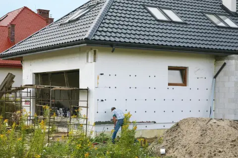 Budowa domu w toku. Izolacja ścian styropianem, widoczny fragment garażu i pracujący mężczyzna. W tle inne domy i stos piasku. Cokół domu jest jeszcze nieotynkowany.