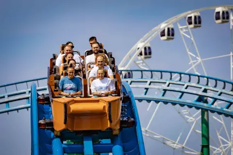 Ludzie na wagoniku rollercoastera przeżywają emocje podczas zjazdu. W tle widać koło widokowe.