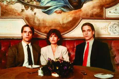 Jeremy Irons w filmach, tutaj z dwiema innymi osobami, siedzi przy stole w eleganckiej restauracji.