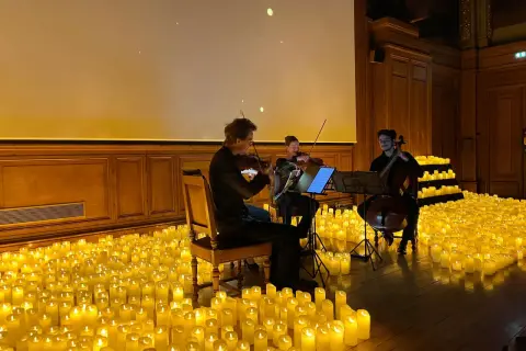 Un trio de musiciens joue dans une salle baignée par la douce lueur de milliers de bougies. Un concert à la bougie magique.
