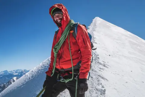 Uśmiechnięty alpinista na szczycie Mont Blanc, w pomarańczowej kurtce i z liną, cieszy się widokiem Alp.