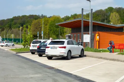 Stationnement sur une aire d'autoroute avec plusieurs voitures garées. Un homme est assis sur un banc rouge devant un bâtiment orange.