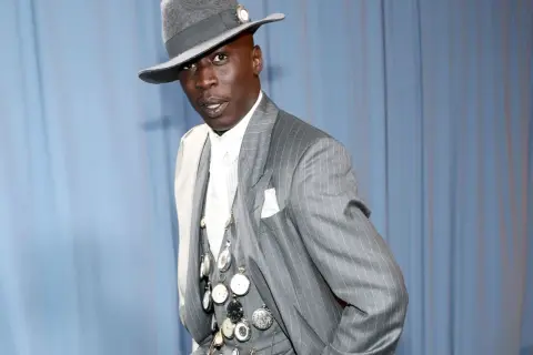 Un uomo con un cappello grigio e un completo gessato, ornato da numerosi orologi da taschino, incarna l'eleganza maschile.
