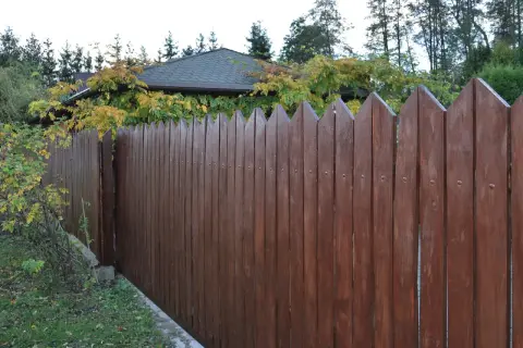 Solidny, brązowy płot z desek z ostro zakończonymi sztachetami, oddzielający ogród od domu z jesiennymi liśćmi.
