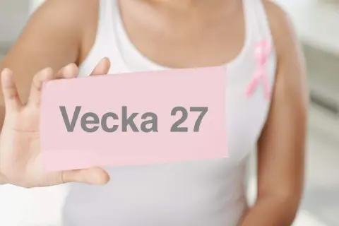 Gravid vecka 27. En person håller upp en rosa lapp med texten "Vecka 27". En rosa band pryder personens vita linne.