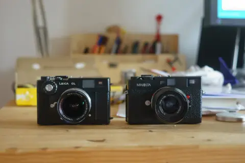 Two classic rangefinder cameras, a Leica CL and a Minolta, sit side-by-side on a wooden desk, ready for their next adventure.
