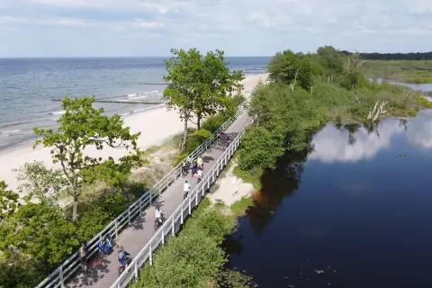Ludzie na rowerach przemierzają malowniczą ścieżkę Velo Baltica, z jednej strony morze i plaża, z drugiej spokojna, ciemna woda.