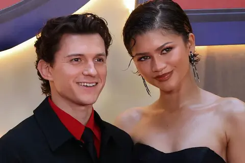 Tom Holland y Zendaya sonríen juntos en un evento. Él lleva una camisa roja y corbata negra. Ella luce un vestido negro y pendientes largos.
