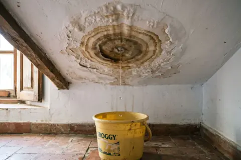 Goteras en el techo, una cubeta amarilla recoge el agua. Urge reparar tejado sin quitar tejas.