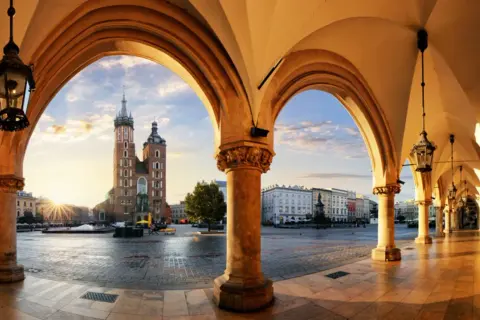 Krakau Geheimtipps: Blick durch Arkaden auf den Marktplatz mit Marienkirche im Sonnenaufgang. Ein malerischer Ort für Entdeckungen.