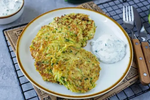 Drei goldbraune Zucchini-Pfannkuchen liegen auf einem Teller, daneben ein Klecks Kräuterquark. Ein leckeres Gericht!