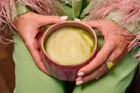 Dłonie trzymają kubek z zieloną matchą, zastanawiając się, czy matcha odchudza. Ubrana w pistacjowy strój z różowymi piórami.