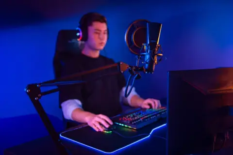 Ein junger Mann mit Kopfhörern sitzt vor einem beleuchteten Mikrofon und einer Tastatur. Er ist ein Twitch Streamer, der seine Spiele oder Inhalte live teilt.