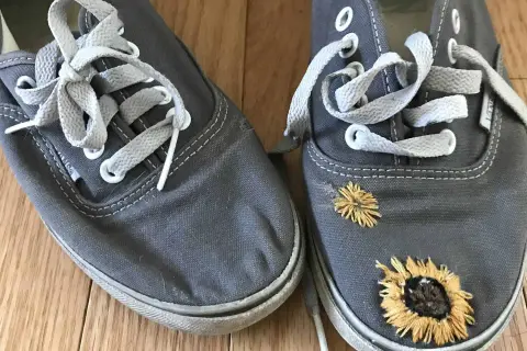 Paire de chaussures grises avec des lacets blancs. L'une des chaussures est ornée de broderies de tournesols, donnant un air bohème à ces chaussures trouées.