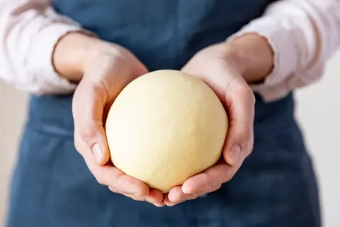 Hände halten einen runden, hellgelben Teigball.