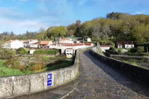 Antiguo puente de piedra con señal de peregrinación. Al fondo, un pueblo con casas de tejados rojos y vegetación exuberante. ¡Ponte Maceira que ver!