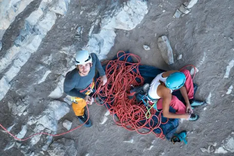 Dwóch wspinaczy przygotowuje się do wspinaczki. Obok nich leży zwinięta, jaskrawoczerwona lina alpinistyczna.