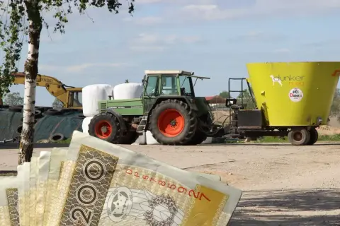 Zielony traktor z przyczepą paszową na tle pola i sterty bel. W tle koparka. Na pierwszym planie polskie banknoty 200 zł. Eudr co to? Inwestycja w rolnictwo.