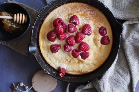 Puszysty omlet z malinami, posypany cukrem pudrem, podany w żeliwnej patelni. Obok miseczka z miodem i plastrem pszczelim.