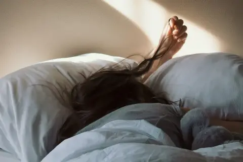 A person sleeps soundly, their hair spread across a pillow. The morning sun casts a warm glow, but the pillow rash is evident.