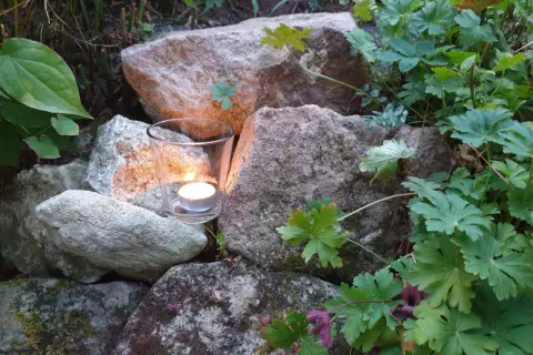 Ein Teelicht in einem Glas leuchtet zwischen Steinen und grünen Pflanzen. Ein Symbol für Hoffnung und einen bibelvers mut.