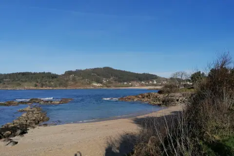 La tranquila playa de Vilariño, con arena dorada y aguas azules, se extiende bajo un cielo despejado. Rocas salpican la orilla, y colinas verdes cubiertas de árboles la rodean.