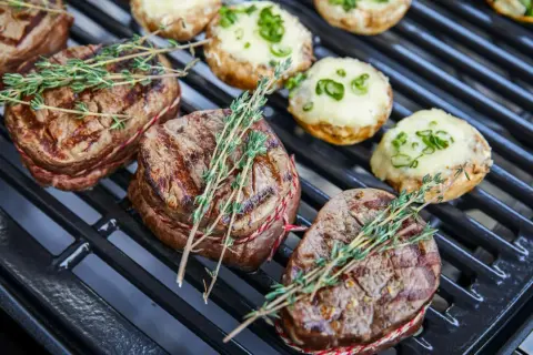 Saftige Rindersteaks mit Kräutern auf dem Holzkohlegrill. Daneben liegen gefüllte Champignons.