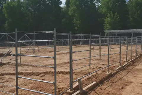 Budowa ogrodzenia z siatki i metalowych słupków na piaszczystym terenie, w tle drzewa.