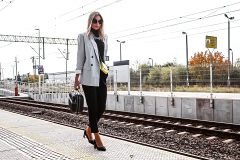 Elegancka szara marynarka damska w stylizacjach na podróż. Kobieta z małą walizką na peronie kolejowym.