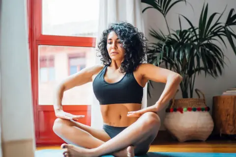 Mujer practicando hipopresivos postparto en casa, sentada en el suelo con las piernas cruzadas.