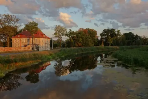 Dworek w Gdańsku Olszynce z czerwonym dachem odbija się w spokojnej wodzie kanału. Chmury na niebie malują zachód słońca.