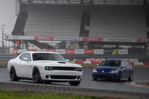 Biały Dodge Challenger i niebieski Volkswagen Golf na torze Słomczyn. W tle trybuny i banery reklamowe.