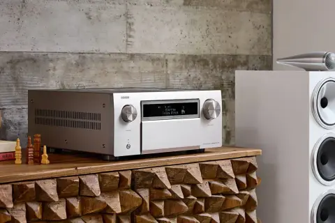 Srebrny amplituner Denon z wyświetlaczem Dolby Atmos, obok białej kolumny głośnikowej. To centrum domowego kina, które pozwala cieszyć się kinową jakością dźwięku.