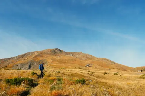 Escursionista in cammino verso il monte più alto dell'Emilia Romagna, con un paesaggio dorato e un cielo azzurro.