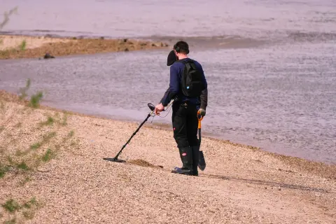 Mężczyzna z wykrywaczem metali i łopatką na plaży przy wodzie.