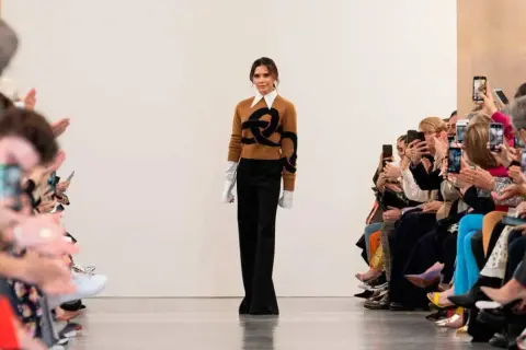 Victoria Beckham går på catwalken, klädd i en brun tröja med ett svart mönster och vida svarta byxor. Publiken, inklusive Harper Beckham, fotograferar med sina mobiler.