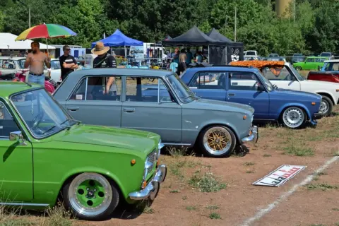 Grüne und graue Lada-Limousinen, ein blauer Lada und weitere Autos stehen auf einem Feld.