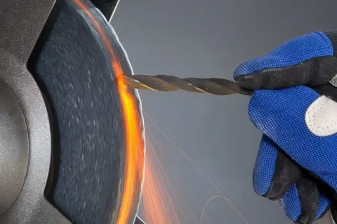Ręka w niebieskiej rękawicy szlifuje wiertło, tworząc iskry. Precyzyjne wiercenie w aluminium wymaga ostrości.