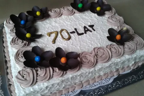 Prostokątny tort z białym kremem, ozdobiony czekoladowymi kwiatami i napisem "70-LAT".