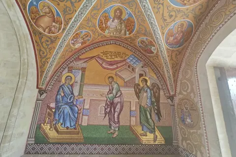 Mozaika przedstawia scenę biblijną z postaciami w stylu sztuki sakralnej. Widać Matkę Boską, anioła i mężczyznę, a także symbole religijne.