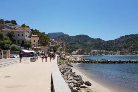Promenad längs stranden i Port de Sóller med folk som njuter av solen och havet, omgivet av gröna berg.