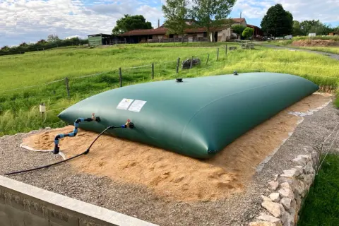 Gran depósito de agua de 20 litros, verde, instalado en un campo con una granja al fondo.