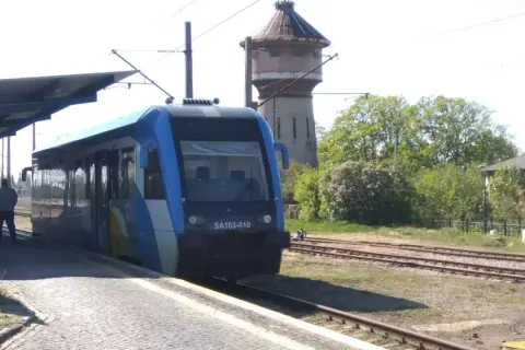 Niebieski pociąg SA103-010 czeka na pasażerów na peronie. W tle stara wieża ciśnień i drzewa. Wygodny wicie dojazd PKP.